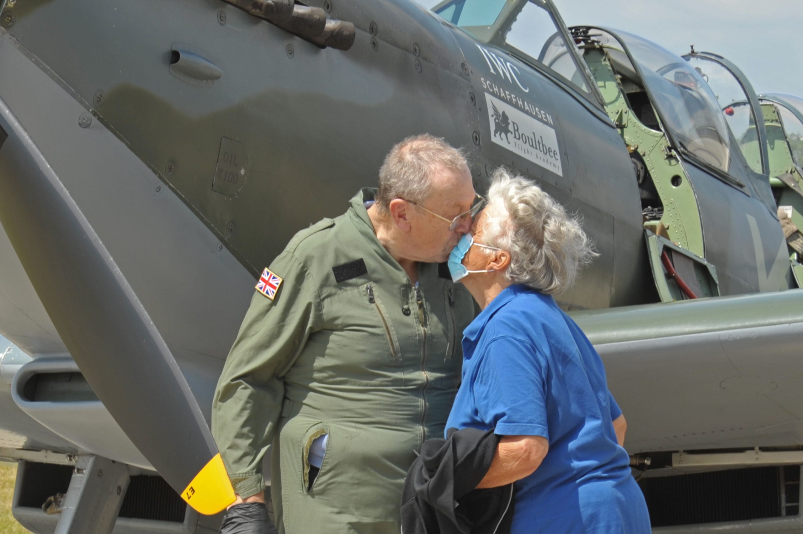 An older couple kissing outside of a spitfire plane.